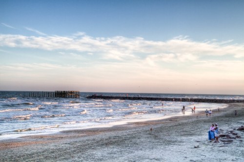 A serene beach scene with gentle waves, a distant pier, and people walking along the shore under a colorful sky.