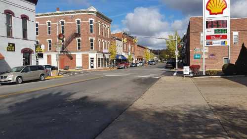 A small town street with brick buildings, shops, and a Shell gas station under a partly cloudy sky.