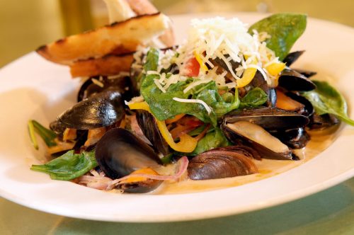 A plate of mussels with spinach, colorful peppers, and grated cheese, served with a slice of toasted bread.