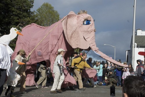 A large pink elephant puppet is paraded by performers, surrounded by a crowd at a festive outdoor event.