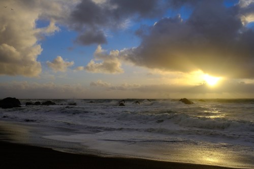 A serene beach scene at sunset, with waves crashing on rocks under a cloudy sky illuminated by golden light.
