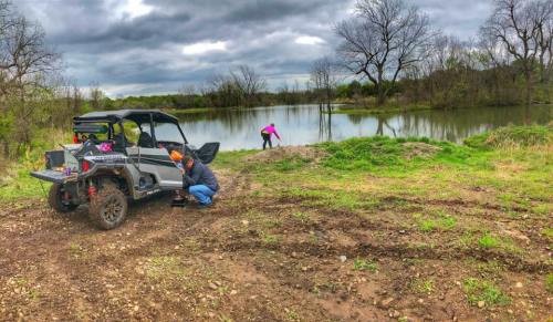 A group near a pond, one person working on an ATV while another explores the shoreline, surrounded by trees and cloudy skies.