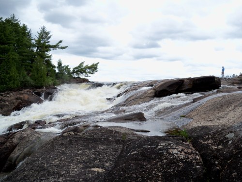 A rushing river cascades over rocky terrain, with a person standing on the shore under a cloudy sky.