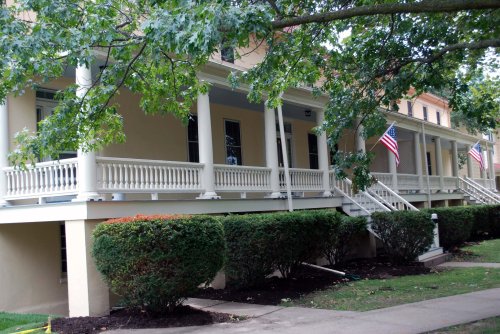 Historic building with white columns and porches, surrounded by green bushes and American flags.