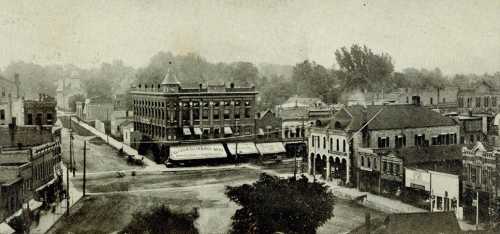 A historic black-and-white view of a town square with buildings, trees, and a street intersection.