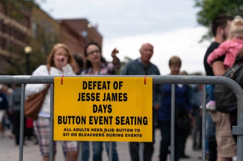 A yellow sign on a barrier reads "Defeat of Jesse James Days Button Event Seating" with a crowd blurred in the background.