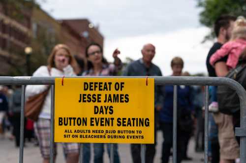 A yellow sign on a barrier reads "Defeat of Jesse James Days Button Event Seating" with a crowd blurred in the background.