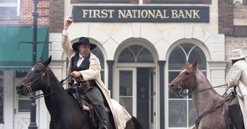 A cowboy on horseback raises a gun in front of a bank, with another horseman beside him.