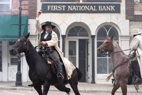 A cowboy on horseback raises a gun in front of a bank, while another rider follows closely behind.