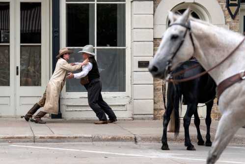 Two men in Western attire appear to be in a playful struggle on a street, with a horse walking by in the foreground.