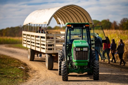 A green tractor pulls a covered wagon along a dirt path, with people walking nearby in a rural setting.