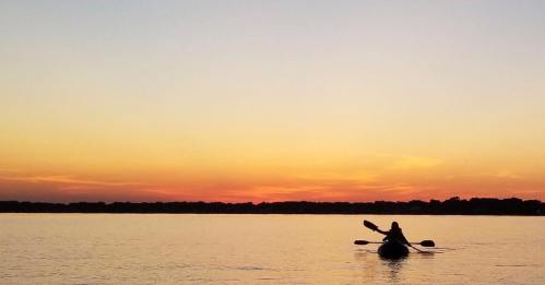 A person kayaking on a calm lake at sunset, with vibrant colors reflecting on the water.