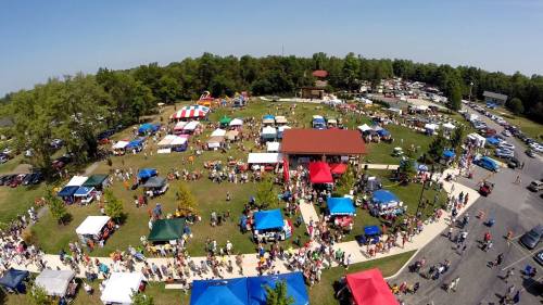 Aerial view of a lively outdoor festival with colorful tents, crowds, and activities in a green park setting.
