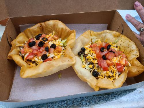 Two taco salad bowls filled with lettuce, cheese, tomatoes, and black olives in a cardboard takeout container.