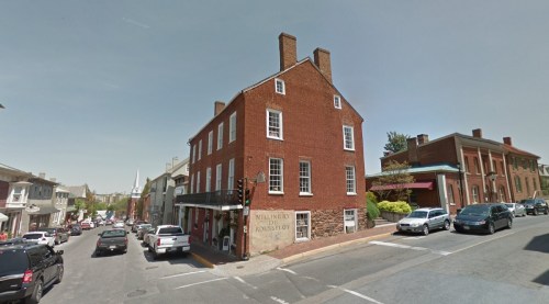 Historic brick building on a street corner, with parked cars and shops nearby in a small town setting.