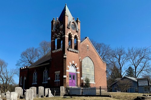 Historic brick church with a tall clock tower, purple doors, and a graveyard in front, set against a clear blue sky.