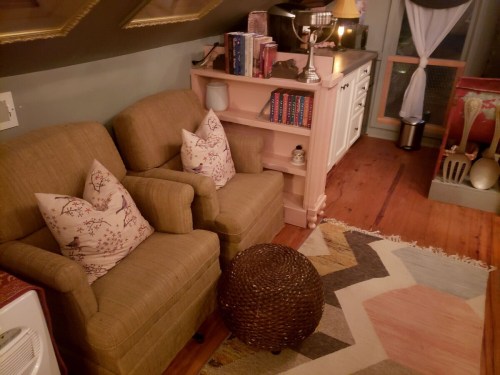 Cozy attic space with two armchairs, a small table, bookshelves, and a colorful rug. Warm, inviting atmosphere.