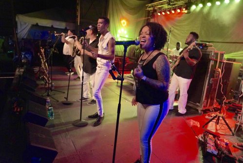 A lively band performs on stage with colorful lights, featuring singers and musicians in white outfits.