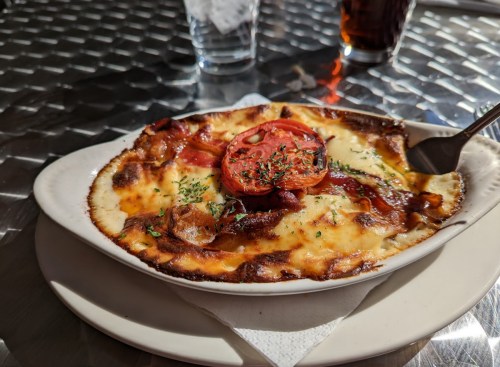 A baked dish with melted cheese, tomatoes, and herbs, served on a plate with a fork, on a metallic table.
