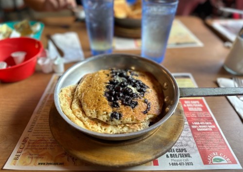 A skillet with blueberry pancakes on a wooden base, surrounded by dining table items and glasses of water.
