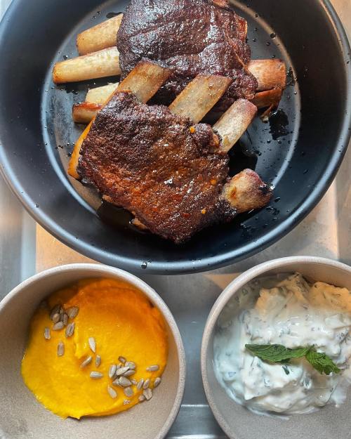 A plate with grilled ribs, accompanied by bowls of creamy sauce and a vibrant orange puree topped with sunflower seeds.