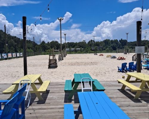 A sandy beach area with colorful picnic tables and volleyball nets under a bright blue sky with fluffy clouds.