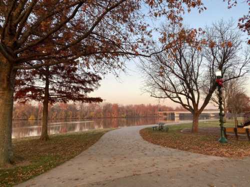 A serene park path lined with trees, leading to a calm lake surrounded by autumn foliage.