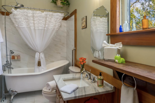 Bright bathroom featuring a vintage tub, white ruffled curtain, wooden accents, and a small vase of orange flowers.