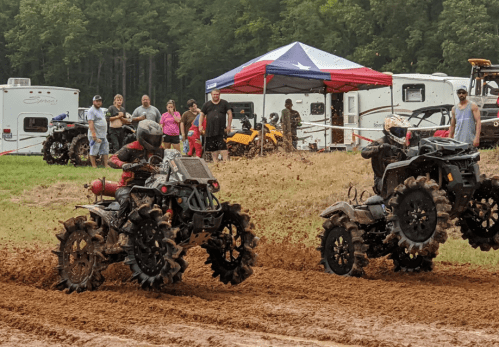 Two ATVs race through muddy terrain, kicking up dirt, with spectators and tents in the background.