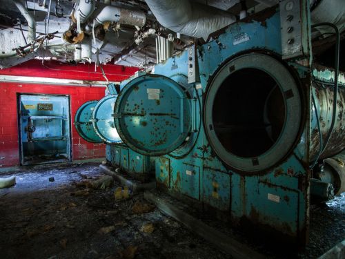 Abandoned industrial laundry room with rusty machines and a red wall, showing signs of neglect and decay.