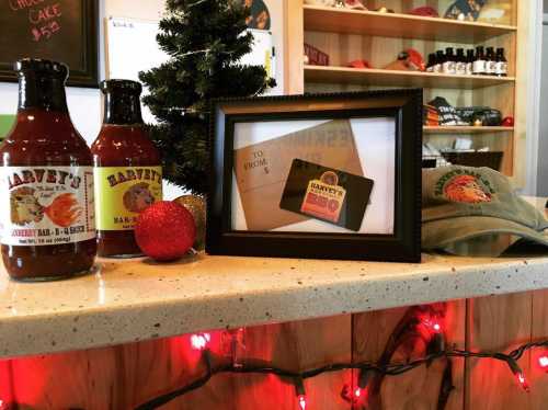 Bottles of BBQ sauce, a gift card, a framed card, and a hat on a counter decorated with holiday lights and a small tree.