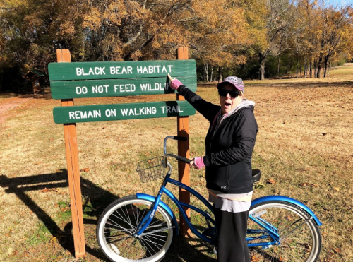 A person in a hat and sunglasses points at a sign that reads "Black Bear Habitat" next to a blue bicycle.