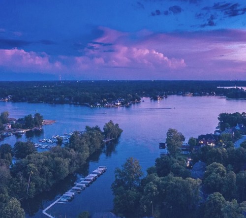 A serene twilight view of a lake surrounded by trees and houses, with boats docked along the shore under a colorful sky.