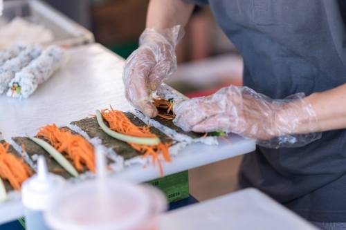 A person in gloves prepares sushi by rolling vegetables and rice in seaweed on a countertop.