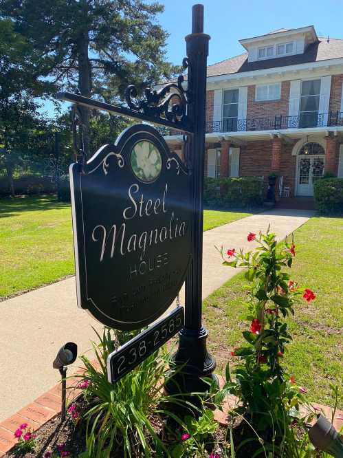 Sign for "Steel Magnolia House" in front of a brick house, surrounded by green grass and flowers.