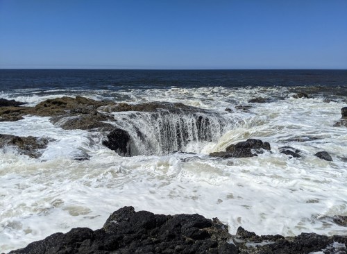 A rocky shoreline with waves crashing against the rocks, creating a small water spout under a clear blue sky.