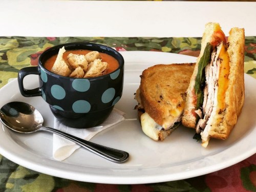 A plate with a grilled sandwich and a cup of tomato soup topped with croutons, set on a colorful tablecloth.