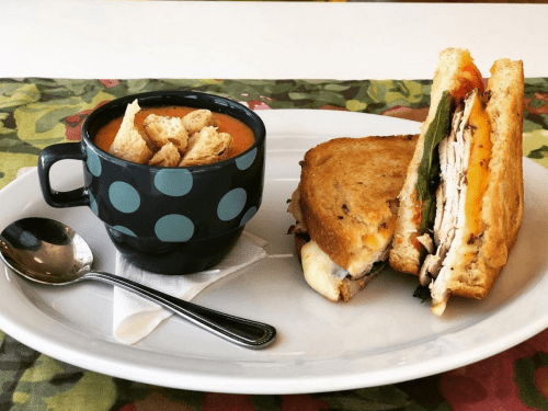 A plate with a grilled sandwich and a cup of tomato soup topped with croutons, set on a colorful tablecloth.