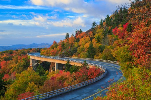 A winding road curves through vibrant autumn foliage under a blue sky with scattered clouds.