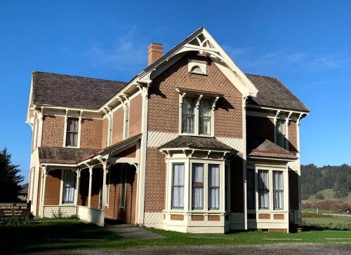 A large, two-story Victorian house with a brown exterior, white trim, and multiple windows under a clear blue sky.