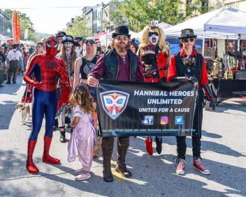 A group of costumed individuals, including Spider-Man, pose with a banner at a street festival.