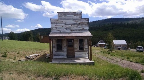 A rustic wooden building stands alone in a grassy area, surrounded by trees and under a blue sky with clouds.