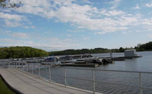 A serene lake scene with docks, boats, and a clear blue sky dotted with clouds, surrounded by lush greenery.