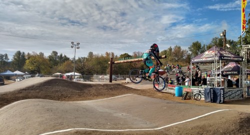 A BMX rider performs a jump on a dirt track, with tents and spectators in the background under a cloudy sky.