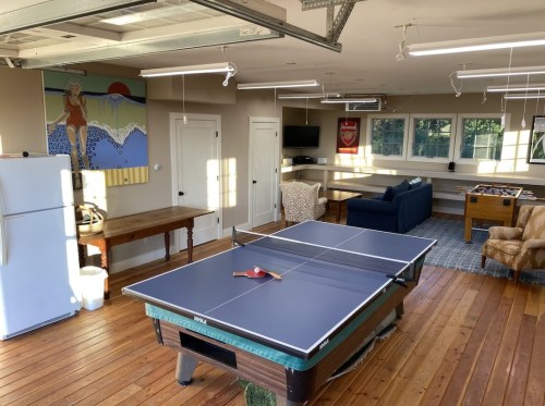 A spacious garage with a ping pong table, seating area, fridge, and artwork on the wall, featuring wooden flooring.