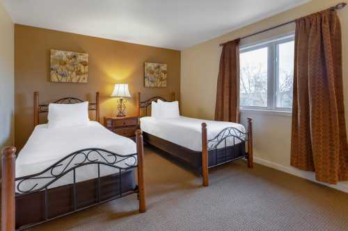 A cozy hotel room with two twin beds, a nightstand, and a window with curtains, featuring warm wall colors.