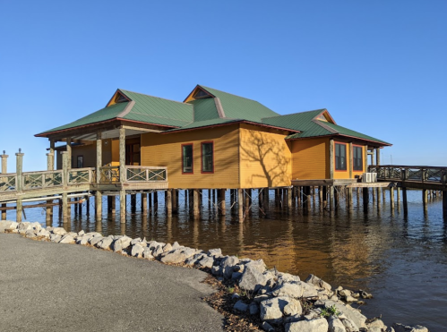 A colorful yellow house on stilts over water, with a green roof and a clear blue sky in the background.
