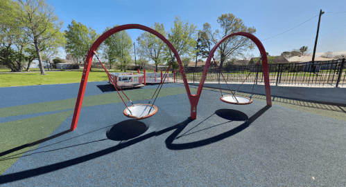 Two red swings on a playground with a blue and green rubber surface, surrounded by trees and a fence.