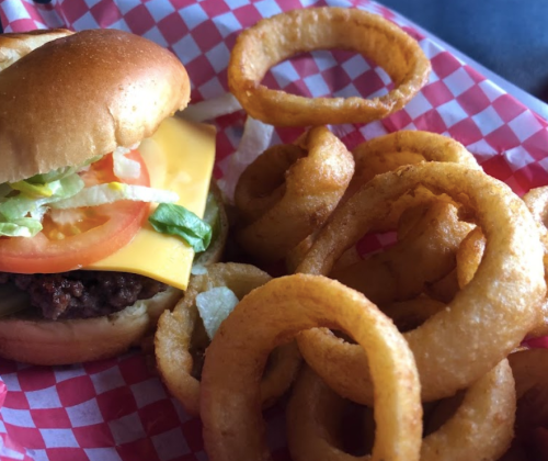 A cheeseburger with lettuce, tomato, and cheese, served with a side of crispy onion rings in a red checkered basket.