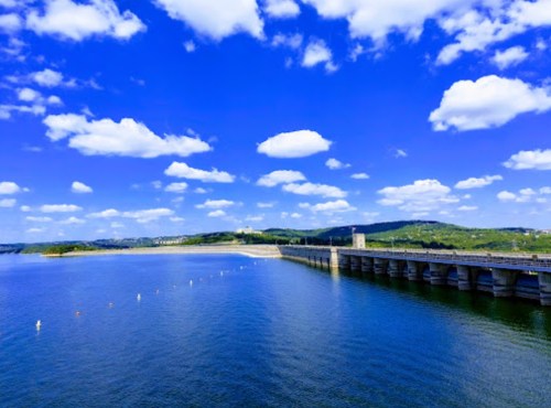 A scenic view of a dam with a blue sky, fluffy clouds, and calm water reflecting the landscape.